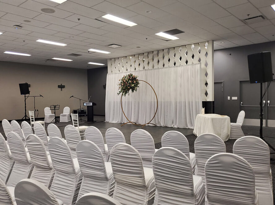 A decorated indoor event space with rows of white covered chairs facing a stage, which features a gold circular arch with flowers, microphones, a podium, and a white-clothed table against a patterned backdrop.