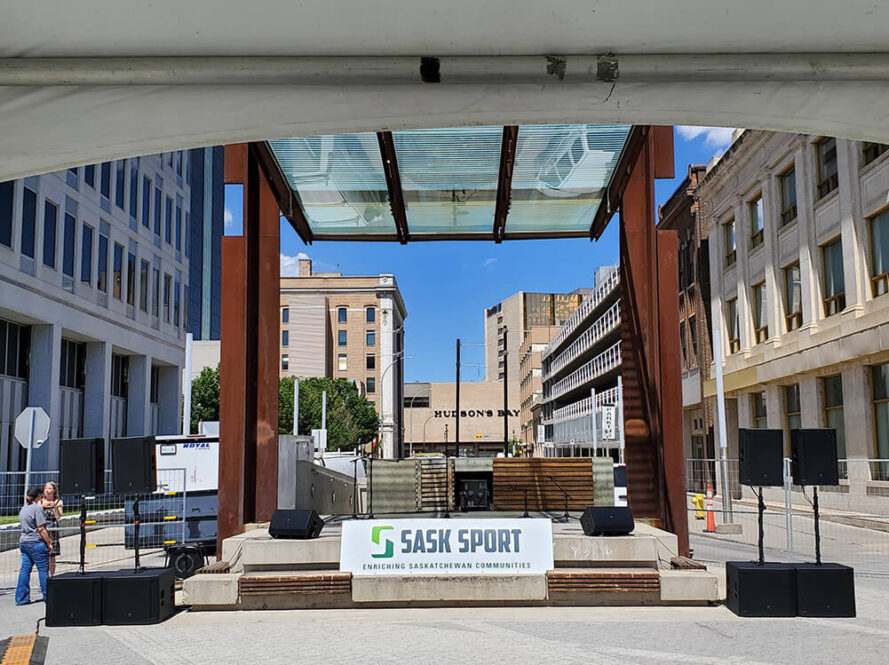 A small outdoor stage with a “Sask Sport” banner is set up in an urban plaza between office buildings, under a glass canopy, with speakers and barricades visible, and a clear blue sky overhead.