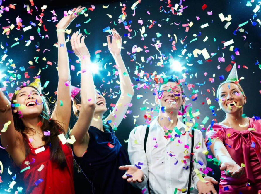 Four people dressed in party clothes celebrate under falling confetti, smiling and raising their hands in the air. Bright lights shine in the background, creating a joyful and festive atmosphere.