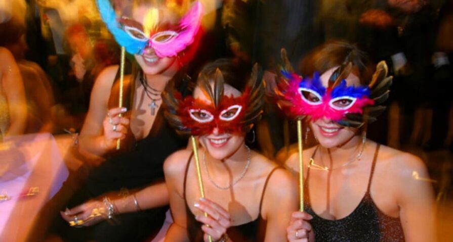 Three women in sparkly dresses sit together at a masquerade party, smiling and holding colorful feathered masks on sticks in front of their faces. The background is blurred, giving a lively atmosphere.