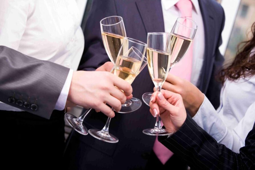 Four people in business attire clink champagne glasses together in a celebratory toast. Only their hands and part of their torsos are visible in the image.
