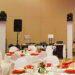 A banquet hall setup with round tables covered in white cloths and red napkins, centerpiece flowers, and a DJ booth with speakers and equipment in the background.