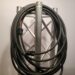 A coiled black power cable is hanging on a metal truss structure. The cable has a plug at one end and is bundled neatly, ready for use or storage. The truss has a crisscross design, and the background is plain white.