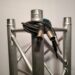 A metal stage truss with a coiled audio cable draped over it, featuring XLR and quarter-inch jack connectors. The truss structure consists of cylindrical tubes connected by diagonal supports, set against a plain background.