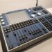 A blue and black lighting control console with multiple buttons, knobs, sliders, and a large touchscreen in the center. The model appears to be a ChamSys MagicQ. Its placed on a white surface.