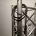 A black cable with connectors is coiled around a silver metal truss structure against a plain background. The truss features a crisscross design, indicating a stage or event setup.