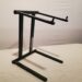 A black metal DJ controller and laptop stand with adjustable arms sits on a white surface. The stand is designed to support equipment at a comfortable height and angle.