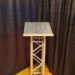 A metal podium stands on a wooden floor with a textured surface and crisscross support design. Dark curtains form the backdrop.