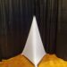 A white, triangular art piece resembling a pyramid stands in front of black curtains on a wooden floor.