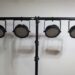 A row of four circular stage lights mounted on a horizontal black pole against a plain white background. Shadows of the lights are cast on the wall behind.