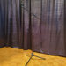 A microphone stand with a boom arm is positioned on a wooden parquet floor in front of dark, textured curtains.