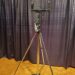 A tall, metal tripod stand with adjustable arms is positioned on a wooden floor. The background features dark, pleated curtains.