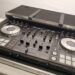 A Pioneer DJ DDJ-SX3 controller is set up on a table. The controller features dual jog wheels, various buttons, sliders, and knobs for mixing music. Its housed in a protective hard case.
