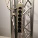 A power strip with six outlets and a power switch is mounted on a metallic truss structure. A plug is attached to the strip, hanging downwards. The background is plain and light-colored.