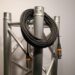 A coiled black cable with connectors is draped over a shiny metal truss structure with cylindrical beams, placed against a plain background.