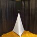 A speaker is mounted atop a tall, triangular white stand against a background of black curtains and wood flooring. The stand resembles a tall pyramid, adding a modern aesthetic to the setting.