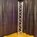 A vertical metal truss stands on a wooden floor, framed by black curtains. The truss has a crisscross pattern and is typically used for supporting lights or speakers in stage setups.