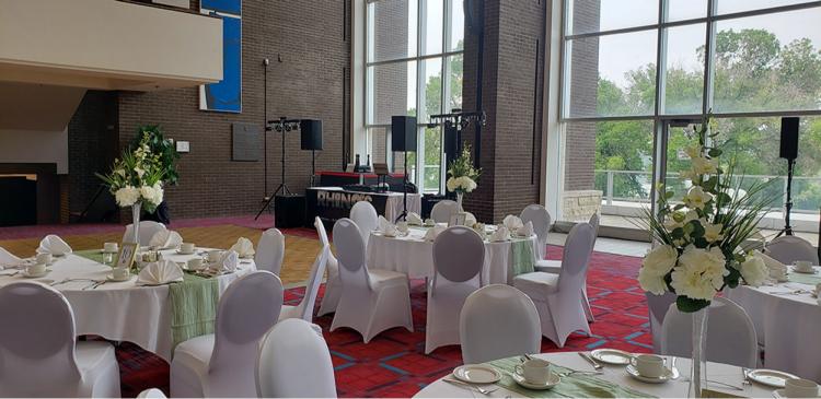 A spacious banquet hall with large windows showcasing a green outdoor view. Tables are elegantly set with white linens, plates, and floral centerpieces. A DJ booth is visible in the background, surrounded by sound equipment.