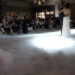 A bride and groom share their enchanting first dance on a cloud-like floor effect, as if they're dancing on a cloud. The room is dimly lit, with seated guests watching in awe.