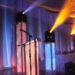 A stage with a clear podium is set against a backdrop of sheer white drapes, illuminated by colorful lights. Two tall pillars with blue lights flank the stage, exuding prestige, while a row of decorated tables hints at an elegant bar/lounge vibe in the background.