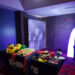 An open air photo booth setup with a table holding various props like masks, hats, and emoji faces, next to a shiny sequin backdrop. A glowing arc light stands nearby, creating a colorful and playful atmosphere in the room.