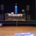 A stage setup for an event features DJ equipment, colorful lights, and a banner reading RHINOs Entertainments and Events. The stage is flanked by tall light towers glowing blue and green. The event space has wooden floors.