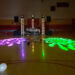A dimly lit gymnasium transforms into the ultimate high school prom venue, featuring a DJ setup with vibrant purple and green stage lights projected onto the wooden floor. Balloons are scattered in the foreground, while string lights twinkle subtly in the background.
