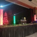 A stage decorated for Christmas features a small tree with red lights, two tall colorful light columns, and a DJ booth labeled RHINOS. This festive setting offers the perfect DJ package experience, with greenery spread across the front under dim lighting.