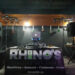The Classic Enhanced Bar/Lounge DJ Package features a DJ booth adorned with a Rhinos Lighting and Sound banner. Speakers and a laptop are poised for performance, with hanging lights adding charm to the dimly lit room’s inviting ambiance.