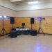 The DJ package is set up in a room with wooden paneling, featuring speakers, light stands, and a table with equipment. A banner reads RHINOS. This classic setup for a high school grad banquet boasts tiled flooring and neutral-colored walls, perfect for any celebration.