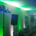 A dimly lit room with green and blue lights showcases a University College DJ setup, featuring speakers, lights, and a laptop on the table. Ready for an electrifying music event, the scene includes a ceiling grid and an emergency exit sign in view.
