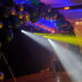 A colorful balloon arch stands as the main event, with blue and yellow spotlights illuminating the area, casting beams of light in a dimly lit room with a draped ceiling, perfect for celebrating a high school grad.