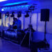 Two DJs stand behind a setup with laptops and sound equipment, surrounded by blue lighting at the high school prom. A banner with DJ Junios is visible on the table. Two large speakers are positioned on either side, and several stage lights are mounted above, setting the perfect mood for a night of dance.