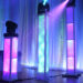Three illuminated pillars with colorful lights emit pink, purple, and blue hues as part of the DJ package, standing on a wooden floor against a backdrop of white draped curtains. Spotlights enhance the ambient atmosphere, creating an electrifying vibe reminiscent of a College DJ event.