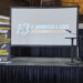 A stage setup with a projector screen displaying E. Bourassa & Sons: Celebrating 100 Years. A podium with a microphone is on the right. A banner on the left lists sponsors and mentions a guest speaker at the event.