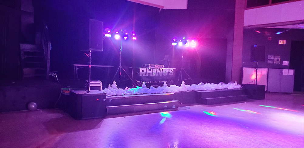 A dimly lit stage with colorful lights and a DJ booth displaying Rhinos in the center. Fog machines line the front, and the dance floor reflects the lights, creating a festive atmosphere.