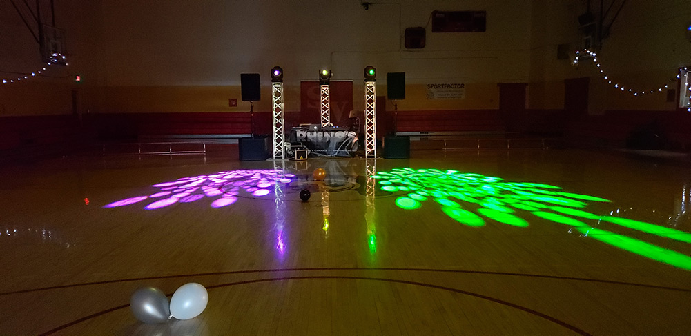 A dimly lit gymnasium with a DJ setup, featuring light towers and colorful spotlights casting patterns on the wooden floor. A few balloons are scattered around, and string lights decorate the upper walls.