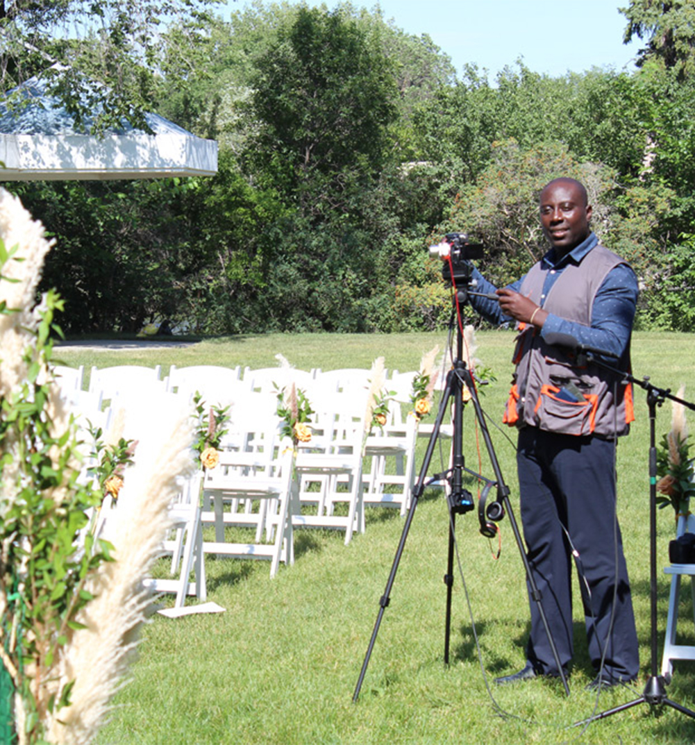 A videographer stands next to a tripod with a camera in a green outdoor setting. Rows of white chairs are arranged for an event, with flowers attached to the aisle seats. Tall grass and trees are visible in the background.
