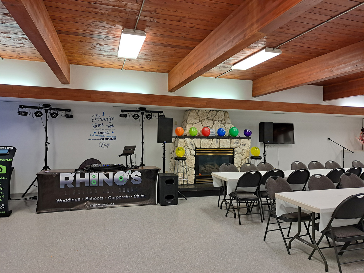 A decorated indoor event space with a DJ booth labeled Rhinos, colorful balloons by a stone fireplace, folding chairs, and tables set up, ready for a gathering or celebration.