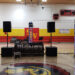 A DJ setup with speakers, lights, and equipment is arranged on a gymnasium floor. Red and yellow bleachers and sports banners are visible in the background, with RHINOS displayed on the DJ table.
