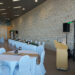 A banquet hall with white tablecloth-covered round tables and a long head table decorated with greenery and lanterns. A wooden podium and audio speakers are set up near a beige stone wall.