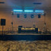A DJ setup on a stage features speakers, lights, and a table with a Rhinos banner advertising events like weddings, schools, corporate, and clubs, in a spacious room with patterned carpet.