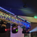 A metal truss with stage lights projects beams of red, green, and blue light in a dark room. An emergency exit sign glows green above a nearby door.