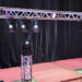 A metal truss lighting rig with spotlights is set up on a stage with red carpeting and a black curtain backdrop. Two smaller trusses support additional lights, and an equipment case sits to the side.