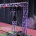 A metal truss structure with stage lights emits colored beams and fog, set up on a red carpet in front of black curtains, likely for an event or performance.