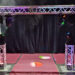 A stage setup features a metal truss arch with colorful spotlights and fog effects, positioned over a red carpet and surrounded by black curtains.