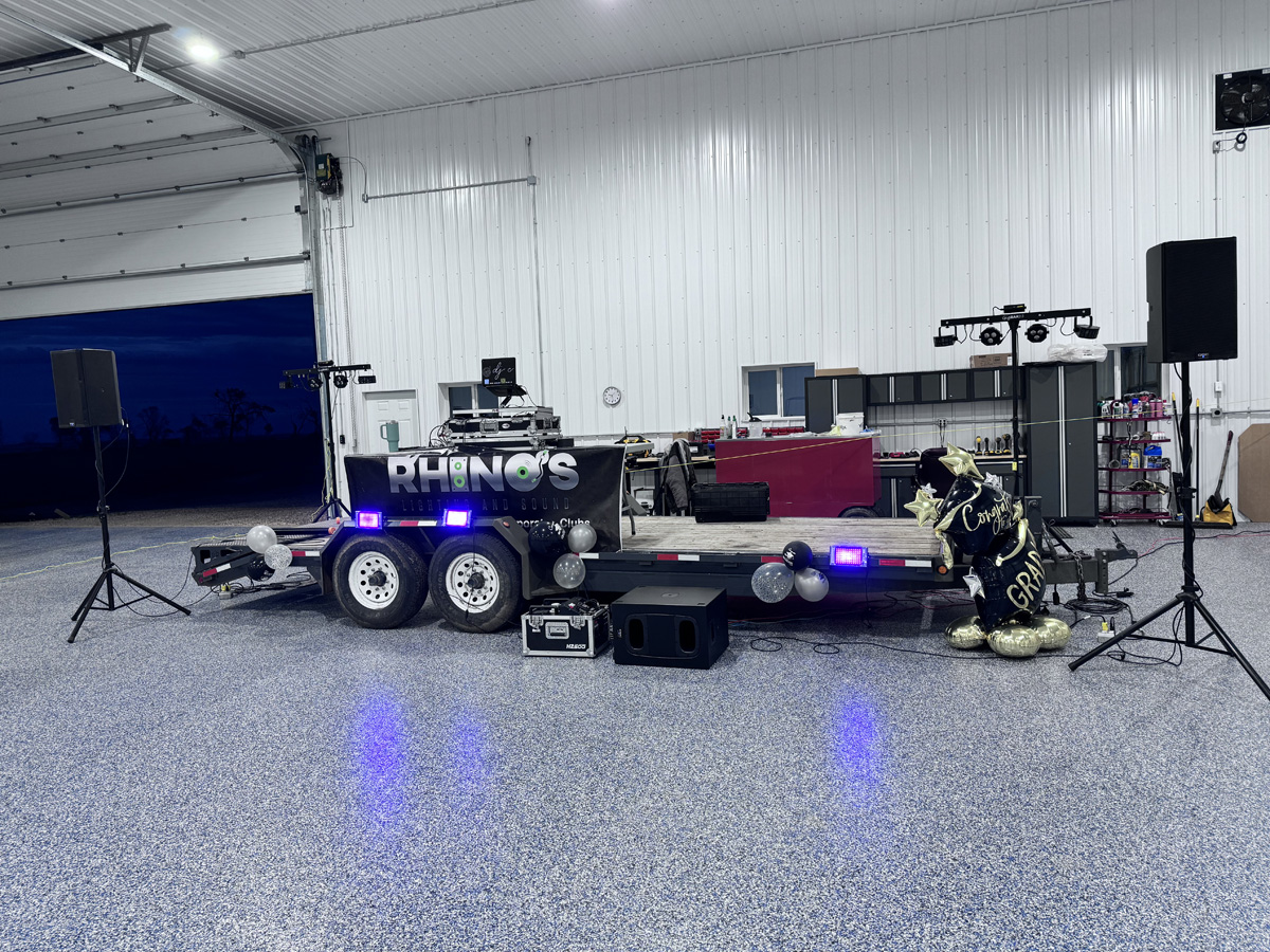 A mobile DJ setup on a trailer with Rhinos written on the side, flanked by speakers and lights inside a spacious garage. Gold and black balloons say Congrats Grad. Equipment and shelves are visible in the background.