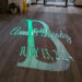 Projected on a wooden floor are the names “Anna & Brody” and the date “JULY 13, 2024” in elegant script, with a large decorative letter “B” in the background at an indoor event venue.