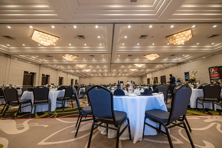 A large, elegant banquet hall with round tables covered in white tablecloths, set for customizing corporate events. Empty chairs surround the tables, and chandeliers hang from the ceiling—perfect for tailored corporate event packages.