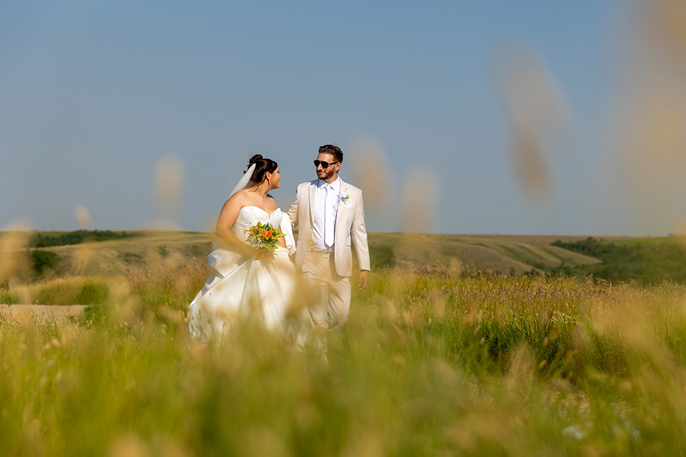 A bride and groom walk together in a sunlit field, smiling at each other, with rolling hills and a clear blue sky in the background—capturing the joy of Greg & Tavia’s Wedding as they celebrate near Lumsden Centennial Hall.